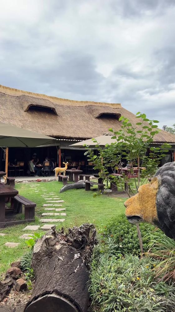 The thatched dining pavilion with guests seated, a stone path, and a crocodile sculpture on the lawn